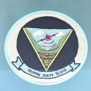 Könnte beinhalten: Gestickter Aufnäher mit schwarzem Hintergrund und einem weißen Dreieck mit blauem Himmel, Wolken und einem roten Flugzeug darin. Das Dreieck ist von einem gelben Rand umgeben. Der Text "OBSRON SIXTY SEVEN" befindet sich auf einem blauen Band unten.