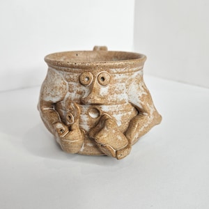 May include: A tan and white speckled ceramic mug featuring a sculpted face with two round eyes, a pointed nose, and a small circular mouth. The mug has two sculpted arms extending from the sides, with one hand holding a small object near the center.