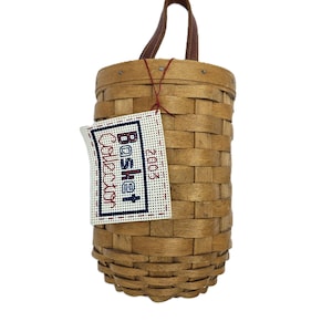 May include: A woven brown basket with a leather handle and a tag that reads "Basket Collection 2003". The basket is cylindrical and has a woven design. The tag is white with blue and red lettering.