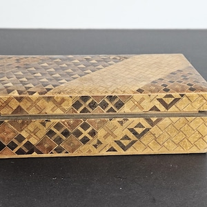 Könnte beinhalten: Rechteckige Holzbox mit geometrischem Muster. Deckel und Seiten zeigen ein Mosaik aus kleinen, kunstvoll angeordneten Formen in Braun- und Beigetönen. Die Box ist geschlossen und liegt auf einer dunklen Oberfläche.