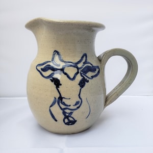 May include: A cream-colored ceramic pitcher with a blue and white cow design. The pitcher has a handle and a spout.