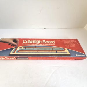 May include: A vintage Cribbage Board game in its original box. The box is red with white and blue accents, and features the words "Cribbage Board" in white. The game board is made of solid hardwood.