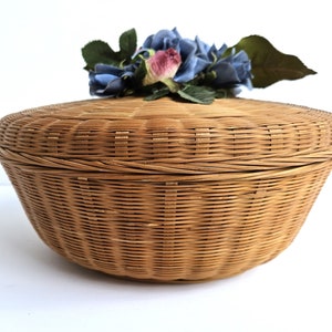 May include: A round wicker basket with a lid, decorated with a cluster of blue and pink fabric flowers. The basket is made of natural woven material and has a light brown color.