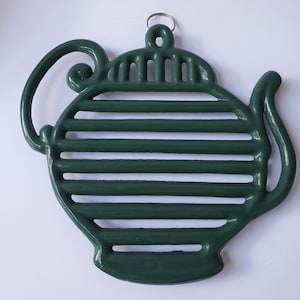 May include: A green cast iron teapot-shaped hot pad with a raised, striped design. The hot pad has a metal loop for hanging.