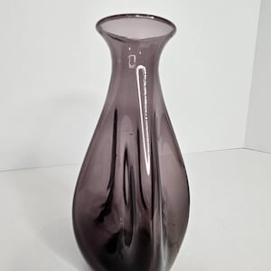 May include: A tall, elegant vase in a deep purple hue. The glass vase has a flared opening and a gently curved body, with subtle vertical ridges adding texture. The vase is empty and sits against a plain white background.
