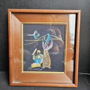May include: A framed embroidery depicting two figures on a black background. The figures are a person holding a blue and green object above their head and a person holding a stick. A blue and green bird is flying in the background.