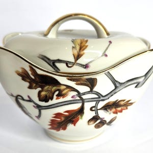 May include: A cream-coloured ceramic serving bowl with a lid. The bowl and lid are decorated with a brown and gold leaf and branch design. The rim and handle are trimmed with gold.