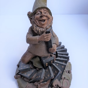 Könnte beinhalten: Eine Keramikfigur eines Zwergs, der einen grünen Hut trägt und einen Stapel Dominosteine hält. Der Zwerg lächelt und hat einen Bart. Die Dominosteine sind zu einem Turm gestapelt und fallen um. Die Figur sitzt auf einem Sockel mit den Worten "FATS" und "1985" darauf.