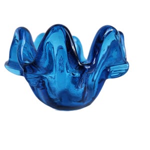 Puede incluir: Un cenicero de vidrio azul vibrante con una forma única y orgánica. El cenicero presenta una serie de curvas onduladas, creando un diseño en forma de pétalo. El vidrio transparente permite el paso de la luz, resaltando el color.