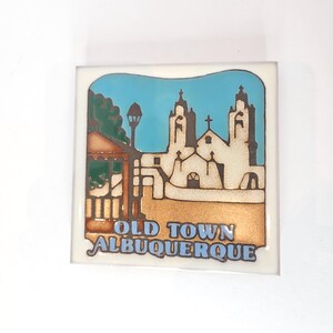 May include: Square ceramic tile featuring a stylized illustration of a church and buildings under a blue sky. The tile has the words "OLD TOWN ALBUQUERQUE" in blue lettering.