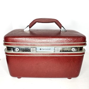 May include: A vintage burgundy Samsonite train case with a matching handle and silver-toned hardware. The case has a rectangular shape with rounded corners and small feet. The Samsonite logo is visible on the front.