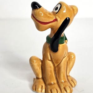 May include: A glossy ceramic figurine of a tan dog with a black nose, black ears, and a green collar. The dog is depicted in a sitting position with its head tilted upward, featuring a wide red mouth and large white eyes with black pupils.