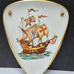 May include: A white ceramic decorative plate shaped like a triangle with rounded edges. The plate has a gold trim and features a hand-painted image of a ship with sails and a red cross. The ship is labeled "Sta Maria".