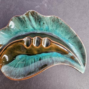 May include: Leaf-shaped ashtray with a glossy finish. The ceramic ashtray features a blend of teal, brown, and black colors. It has three cigarette rests and a textured surface. A decorative item for smokers.