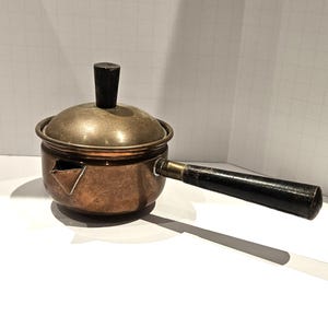 May include: A small copper saucepan with a rounded body, a short pouring spout, and a brass lid featuring a dark wooden knob. The pan includes a long, straight black wooden handle attached with a brass ferrule, showing a metallic finish.