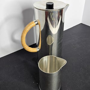 以下が含まれることがあります： 織りハンドルと小さなステンレス製のクリーマーが付いたステンレス鋼のコーヒープレス。コーヒープレスには、蓋に黒いノブが付いています。クリーマーには注ぎ口が付いています。アイテムは黒い表面にあります。