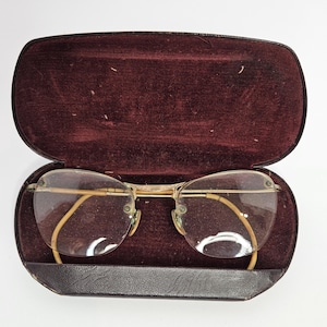 May include: A pair of gold-tone metal eyeglasses with clear lenses and curved earpieces, resting inside an open, oval-shaped, dark brown hard-shell case featuring a deep burgundy velvet interior lining and a hinged lid design.