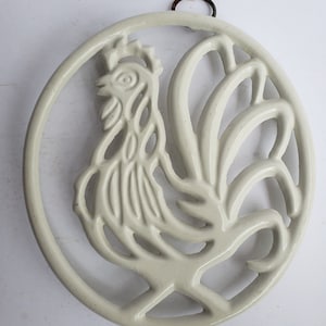 Puede incluir: Salvamanteles de cerámica blanca en forma de gallo con un lazo para colgar. El gallo está de pie con las plumas de la cola extendidas.