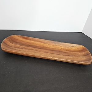 Puede incluir: Una bandeja de madera rectangular y alargada con bordes redondeados. La bandeja es de madera de color marrón claro con vetas visibles. La bandeja está vacía y se encuentra sobre una superficie negra.
