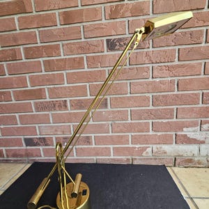 May include: A vintage brass desk lamp with an adjustable arm and rectangular light head. The lamp has a cylindrical base and a switch. The lamp is on a black surface with a brick wall in the background.