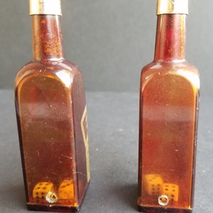May include: Two amber glass bottles with gold tops and dice inside. The bottles are shaped like liquor bottles and have a vintage look.