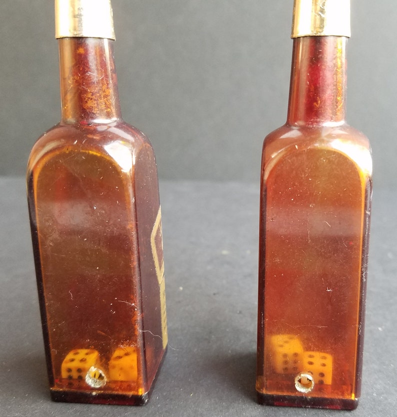 May include: Two amber glass bottles with gold tops and dice inside. The bottles are shaped like liquor bottles and have a vintage look.