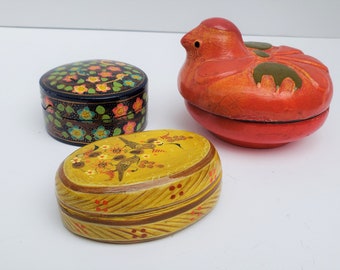 May include: Three hand-painted wooden boxes. One box is round with a floral design in shades of blue, pink, and green. Another box is oval with a yellow background and a floral design with birds. The third box is shaped like a bird and is painted red with green accents.