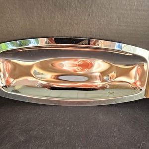 Puede incluir: Una bandeja rectangular de acero inoxidable con asas de madera. La bandeja tiene una superficie pulida y reflectante y bordes curvos. Las asas de madera están unidas con pequeños remaches metálicos y presentan ranuras decorativas.