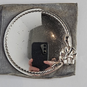 May include: A circular, silver-coloured compact mirror with a decorative floral accent. The mirror has a braided edge and is set in a grey felt pouch. The mirror reflects the surrounding environment.