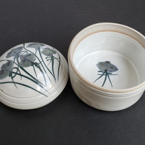 May include: White ceramic lidded jar with a floral design. The lid has a green and purple floral pattern. The jar has a single purple flower with green stems.