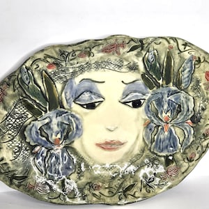 Könnte beinhalten: Ovale dekorative Keramikplatte mit einem Gesicht und zwei blauen Irisblüten. Das Gesicht hat blauen Lidschatten und einen neutralen Ausdruck. Der Hintergrund ist grün und grau mit roten floralen Akzenten.