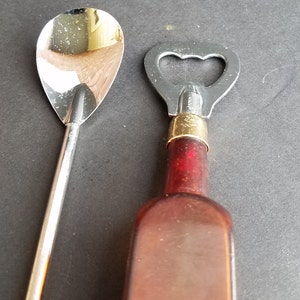 May include: A silver bar spoon and a bottle opener with a gold top and a brown glass bottle.