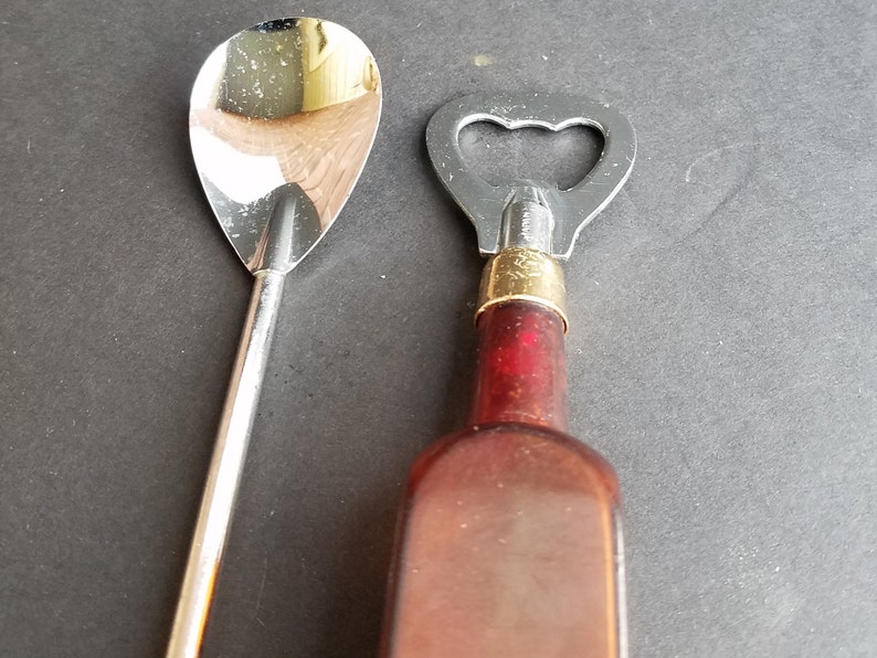 May include: A silver bar spoon and a bottle opener with a gold top and a brown glass bottle.