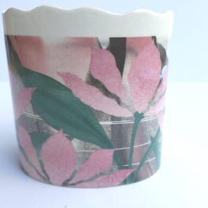 May include: A cylindrical ceramic planter with a scalloped rim. The planter features a floral design with large pink flowers, green leaves, and a gray and white background. The rim is white.