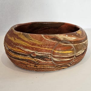 May include: A round, decorative bowl with a textured, layered design. The bowl features bands of brown, yellow, and white, creating a marbled effect. The interior is a darker brown. A unique piece for home decor.