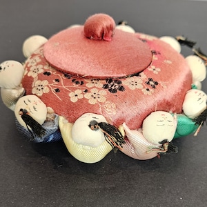 Puede incluir: Un recipiente redondo vintage de tela rosa con tapa y un pequeño botón redondo. El recipiente está decorado con un estampado floral y rodeado de pequeñas figuras blancas con forma de muñeca, con cabello negro y caras pintadas. Las figuras están unidas al recipiente con cintas de colores.