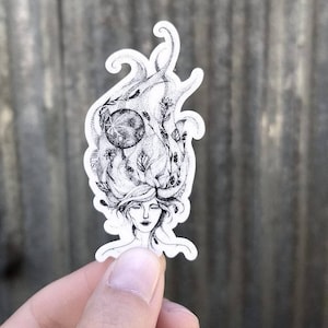 May include: A black and white sticker depicting a woman's face with closed eyes and flowing hair. The hair is decorated with a moon, leaves, and flowers. The sticker has a white border and is held by a hand.