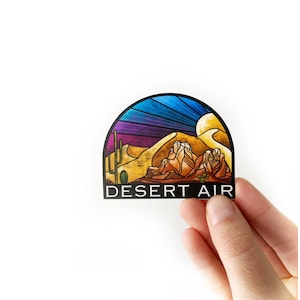 May include: A sticker featuring a desert landscape design. The sticker depicts a sunset with a purple and blue sky, a yellow sand dune, and a mountain range. The text "DESERT AIR" is printed in black at the bottom of the sticker.