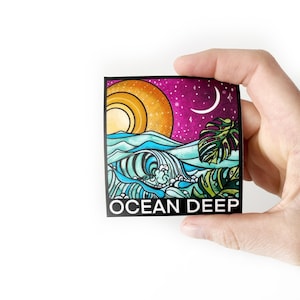 May include: Square sticker with an ocean-themed illustration. The design features a stylized sun, waves, a monstera leaf, and the text "OCEAN DEEP" against a purple starry sky. The sticker is held by a hand.