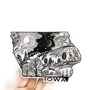 May include: Black and white sticker of the state of Iowa with a detailed design featuring a sun, moon, stars, trees, and the text "IOWA".
