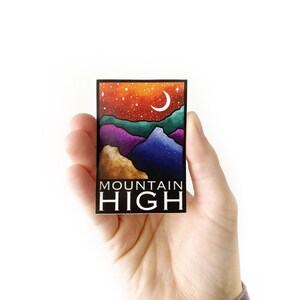 May include: A rectangular sticker depicting a mountain scene beneath a starry night sky and a crescent moon. The colour scheme includes orange, teal, purple, and gold. The text "MOUNTAIN HIGH" is printed in white at the bottom.