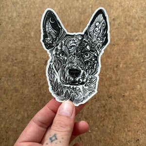 K&ouml;nnte beinhalten: Schwarz-wei&szlig;er Aufkleber mit dem Kopf eines Hundes mit komplizierten Linienkunstdesigns. Der Kopf des Hundes ist mit naturinspirierten Mustern gef&uuml;llt, darunter B&auml;ume, Berge und Sterne. Der Aufkleber befindet sich auf einem wei&szlig;en Hintergrund.