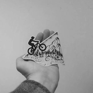 May include: A monochrome sticker depicting a mountain biker on a mountain. The sticker, held in a hand, shows a detailed design of trees and a mountain range, with the biker in silhouette.