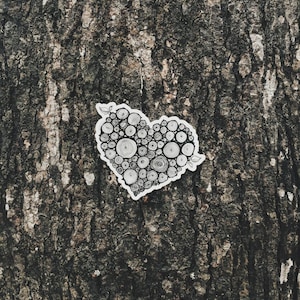 May include: A heart-shaped sticker with a white border, filled with a black and white circular pattern. The sticker is placed on a textured, dark brown tree bark background. The design includes small leaf accents.