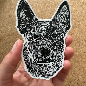 K&ouml;nnte beinhalten: Schwarz-wei&szlig;er Aufkleber mit dem Kopf eines Hundes, der mit einem detaillierten Design von Naturelementen wie B&auml;umen, Bergen und Wolken verziert ist. Der Aufkleber tr&auml;gt den Text "WILD SLICE" am unteren Rand.
