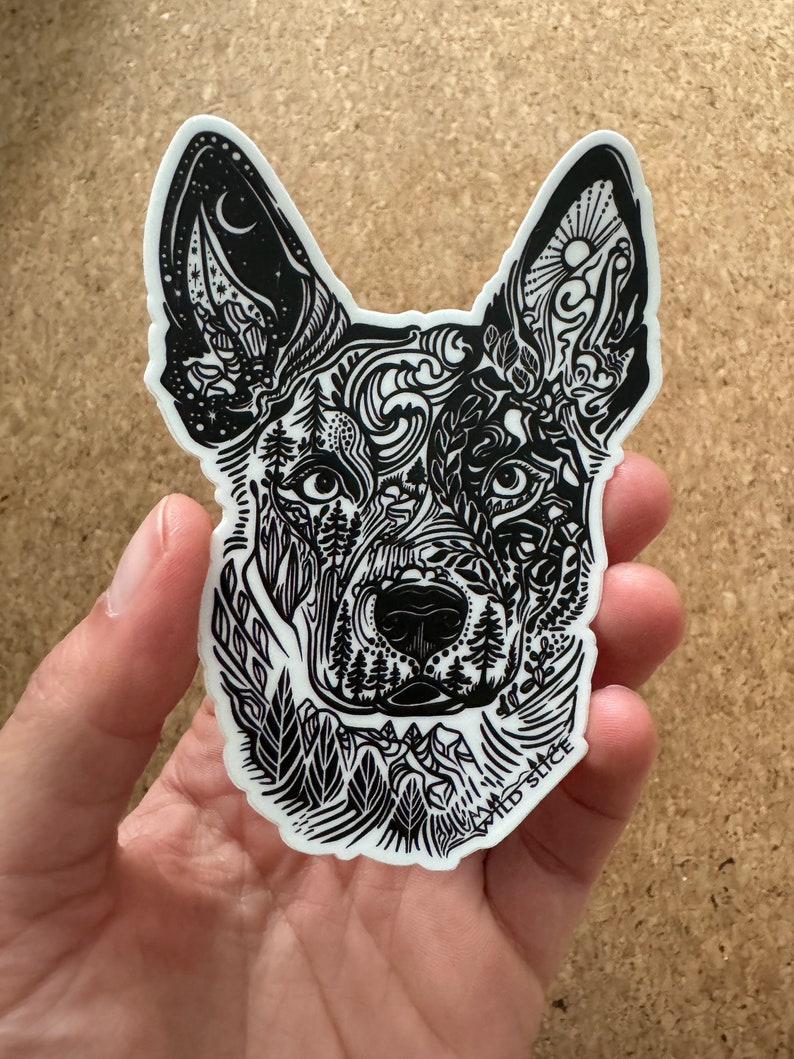K&ouml;nnte beinhalten: Schwarz-wei&szlig;er Aufkleber mit dem Kopf eines Hundes, der mit einem detaillierten Design von Naturelementen wie B&auml;umen, Bergen und Wolken verziert ist. Der Aufkleber tr&auml;gt den Text "WILD SLICE" am unteren Rand.