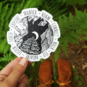 May include: A white round sticker with black ink depicting a stylised tree with a crescent moon and a planet in the night sky. The sticker has text around the edge that reads "FLY ~ BREATHE ~ LISTEN ~ TOUCH ~ EXPLORE ~ WANDER ~ ROAM ~ CONNECT ~ WALK".