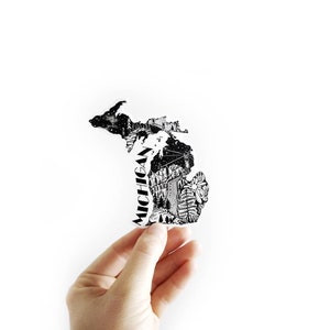 May include: A black and white sticker in the shape of the state of Michigan, held by a hand. The sticker features a detailed illustration of the state's landscape, including trees, a waterfall, and the word "MICHIGAN" in a vertical font.