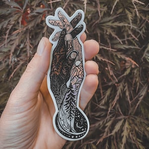 May include: A black and white sticker featuring a detailed illustration of two hands clasped together. The hands are intertwined with nature-inspired designs, including trees, mountains, and stars. The sticker is held in a hand against a blurred background of green foliage.