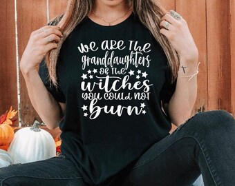 May include: A black t-shirt with white text that reads "We are the granddaughters of the witches you could not burn." The text is surrounded by white stars.